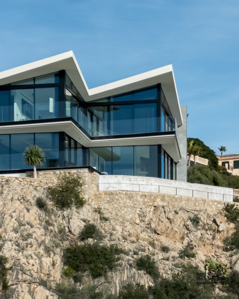 Modern two-story villa with sharp angular roofs and large glass walls built into a rocky hillside under a blue sky.