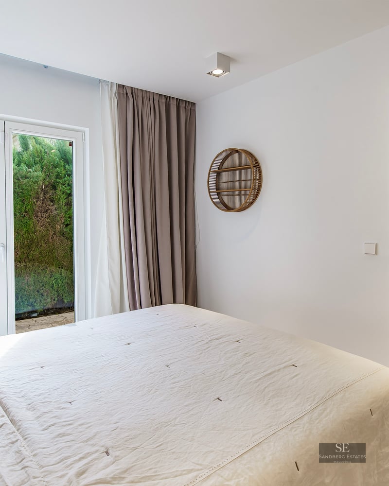 Bright master bedroom with white linens, glass doors to a garden, and a view into an ensuite bathroom.