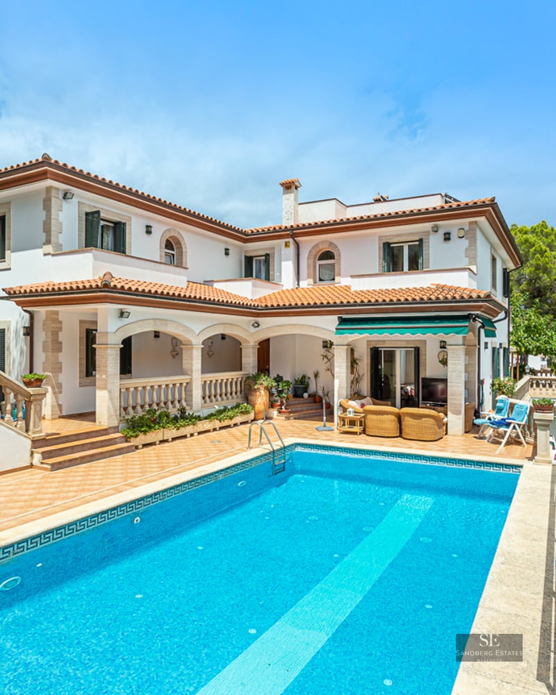 Large rectangular swimming pool in front of a white two-story Mediterranean villa with terracotta roof and stone arches.