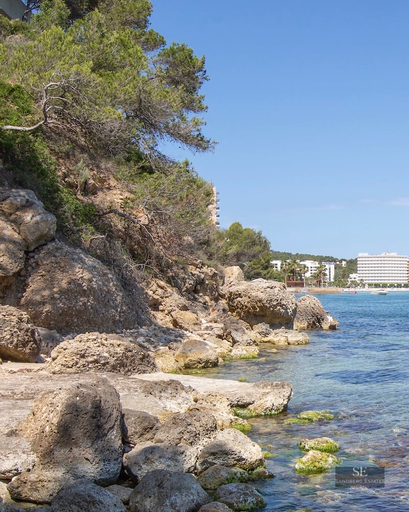 Stone stairs leading down a rocky cliff to turquoise sea with boats and buildings in the distance.