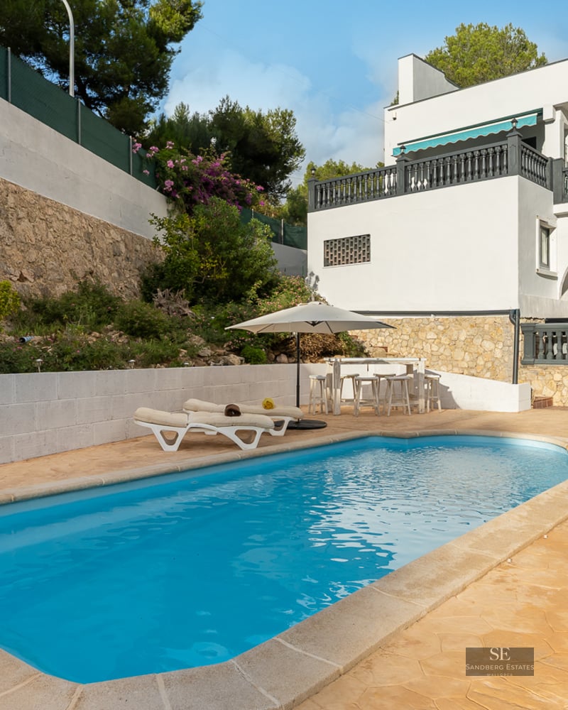 Piscine rectangulaire à l'eau turquoise entourée d'une terrasse en pierre, de chaises longues et d'une villa blanche.