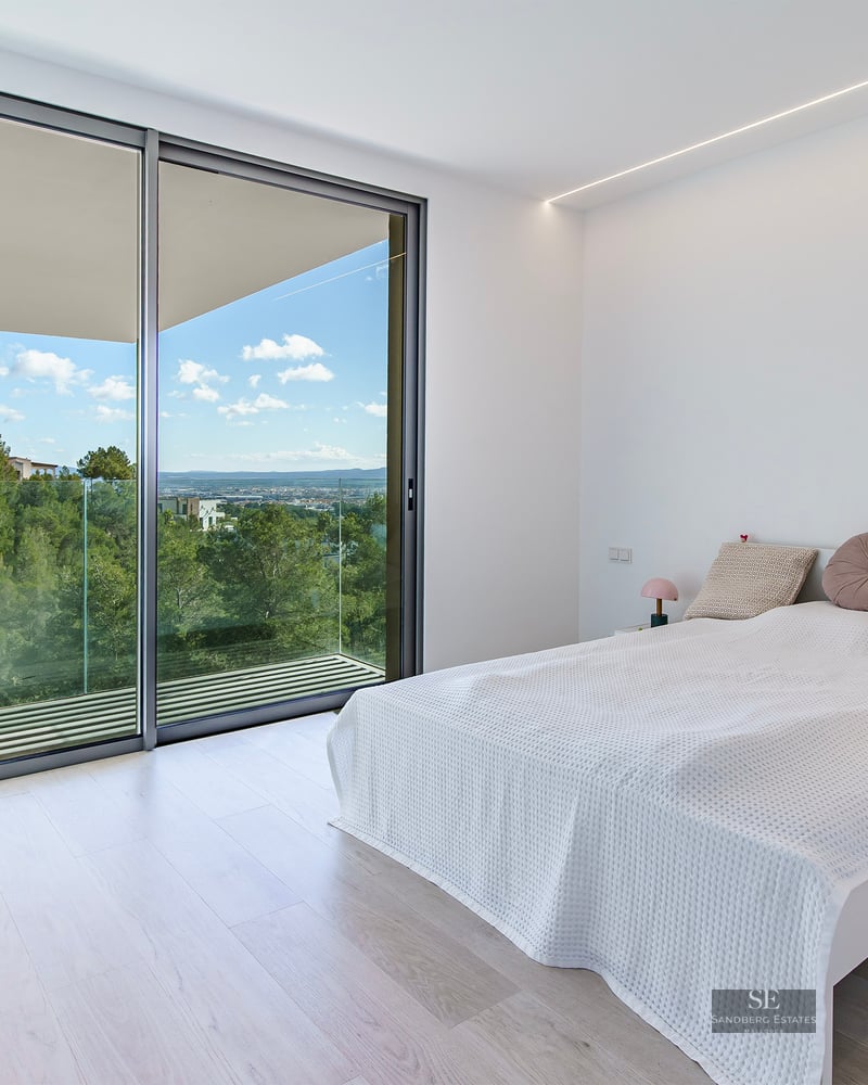 Bright minimalist bedroom with large glass sliding doors opening to a balcony with views of green hills and a blue sky.