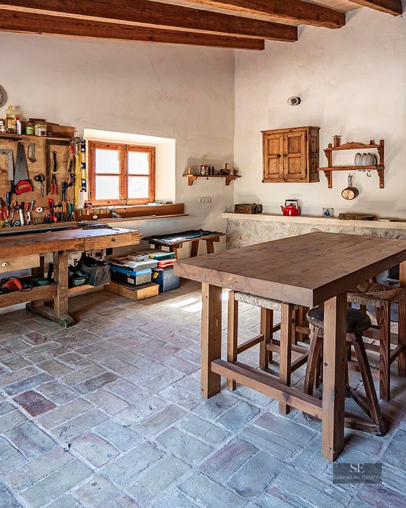 A traditional workshop with a large wooden workbench, tools on the wall, and rustic stone tile floors.