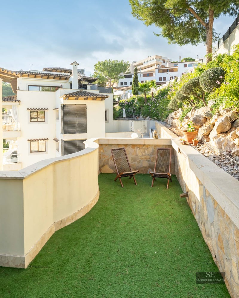 Sunlit terrace with green artificial grass, two wooden lounge chairs, and a natural stone wall.