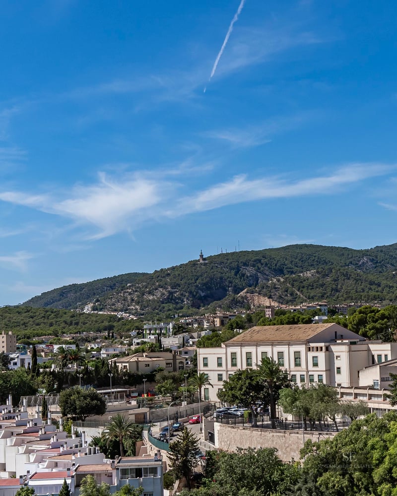High-angle view of white houses in a Mediterranean town with lush green mountains and a blue sky in the background.