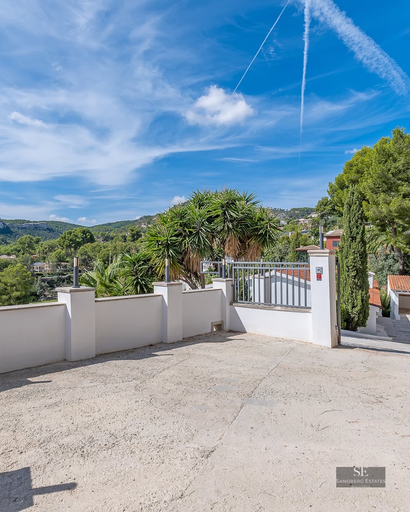 A wide concrete driveway surrounded by white walls and palm trees, leading to a garage with mountain views in the distance.