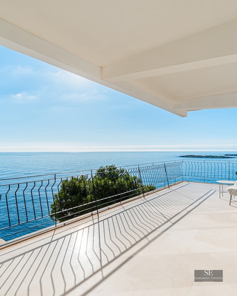 Wide modern terrace with stone flooring and black railing overlooking a calm blue sea under a bright sky.