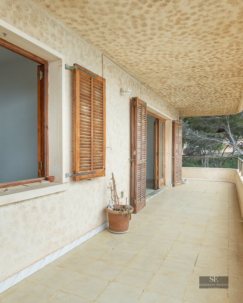 A long balcony with beige tiles, wooden shutters, and a metal railing overlooking lush green trees.