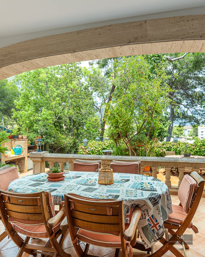 A covered stone terrace with a round dining table, wooden chairs, and a stone balustrade overlooking a lush garden.