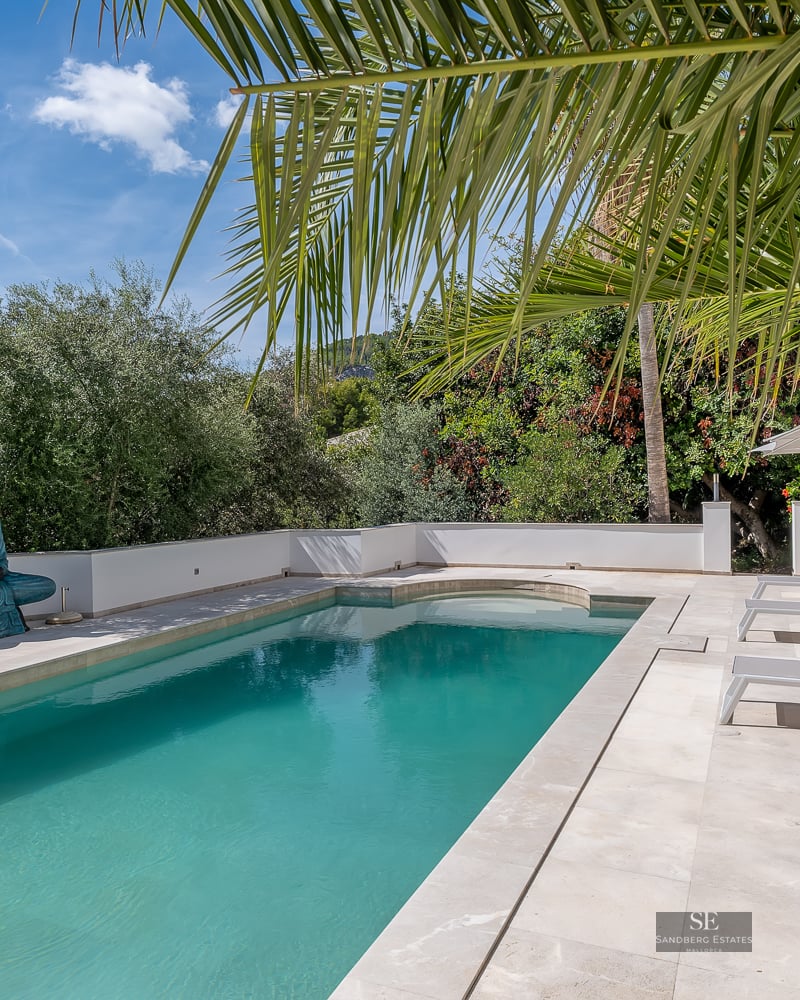 Turquoise pool with white stone tiling, lounge chairs, and a Buddha statue under palm trees.