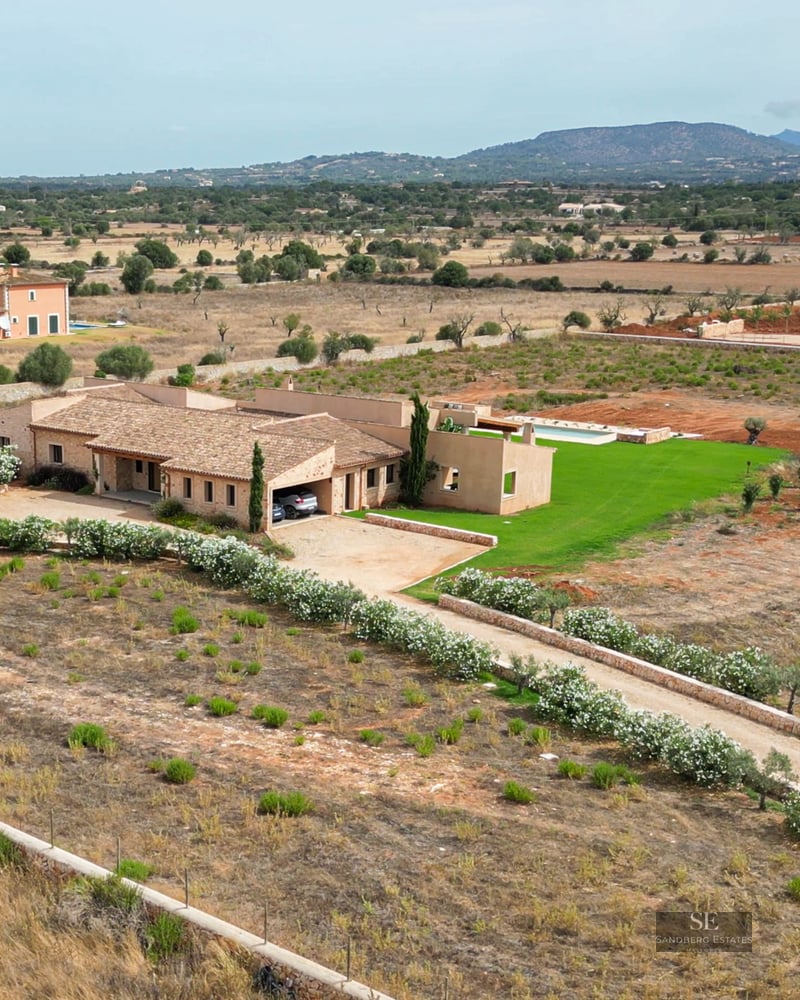 Aerial shot of a stone villa with a pool and flower-lined driveway in a Mediterranean countryside setting.