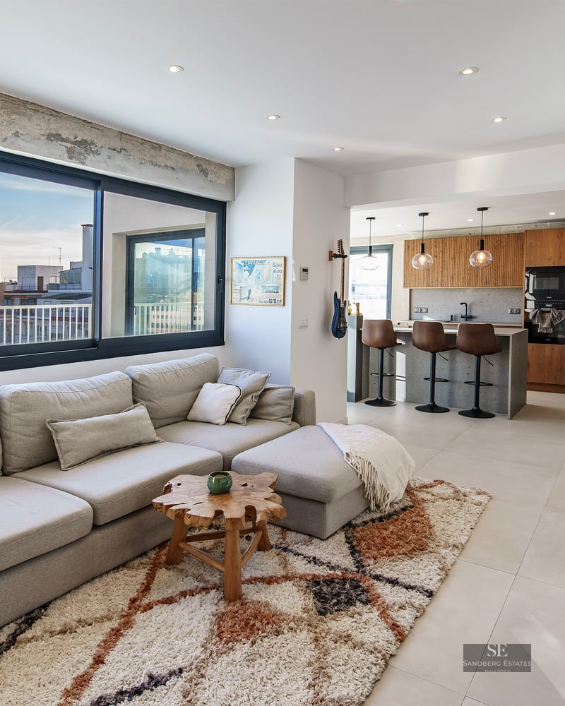 Bright modern living room with a large gray sectional sofa, textured rug, and an open kitchen with city views.