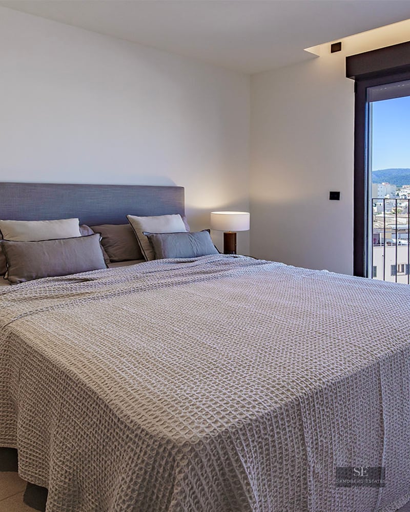 Modern bedroom featuring a large bed with grey bedding, stylish lamps, and a glass door overlooking a city and mountains.