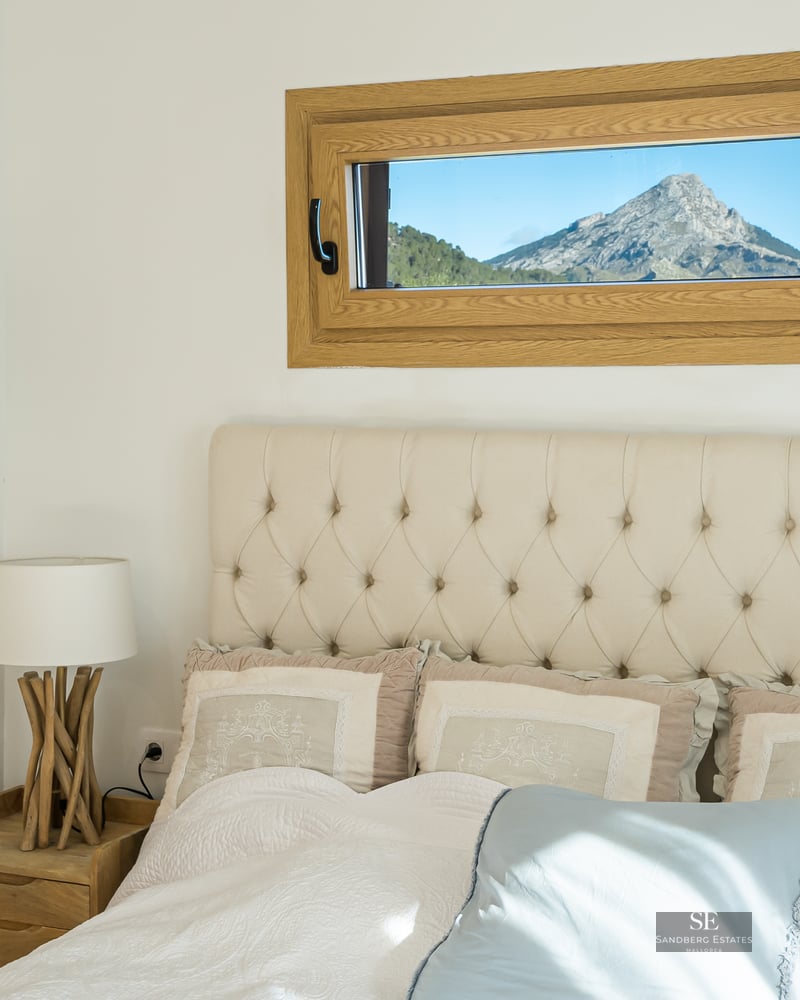 Bed with beige tufted headboard next to a small horizontal window framing a rugged mountain peak.