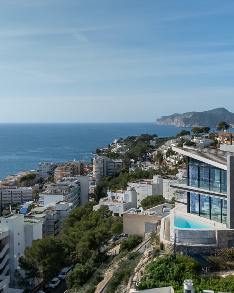Modern white villa on a green hillside with a small infinity pool overlooking a coastal town and the blue sea.