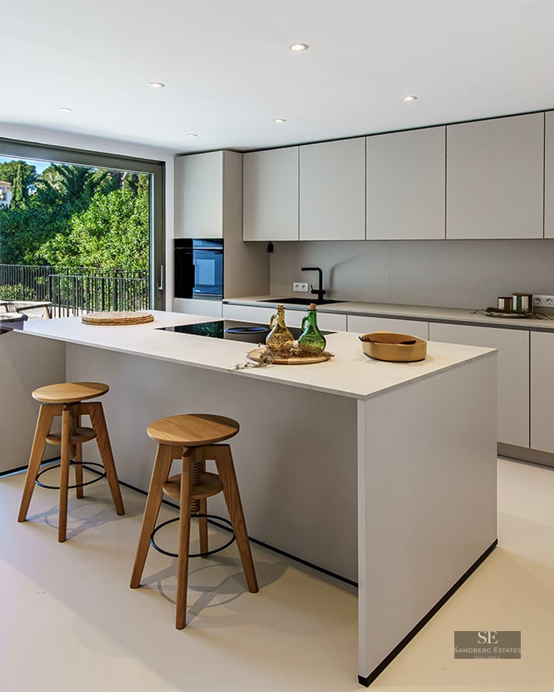 Modern white kitchen with central island, two wooden stools, and large glass sliding doors opening to a terrace and pool.