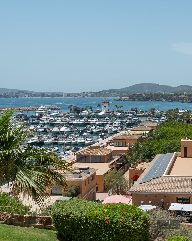 Elevated view of a Mediterranean marina filled with luxury yachts, overlooking the sea and coastline under a clear sky.