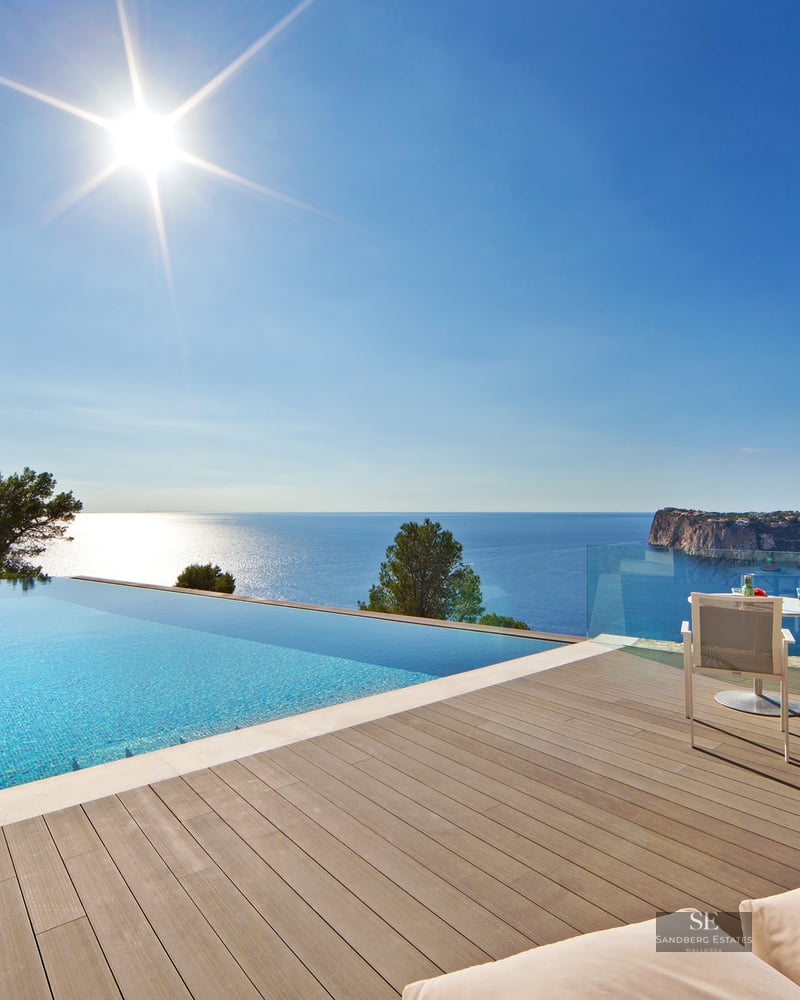 Infinity pool with turquoise water on a wooden deck overlooking a blue sea and rugged coastline under a bright sun.