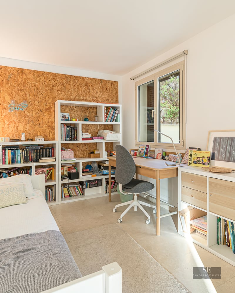 Child's bedroom featuring a white bed, wooden desk, and extensive shelving filled with books under a bright window.