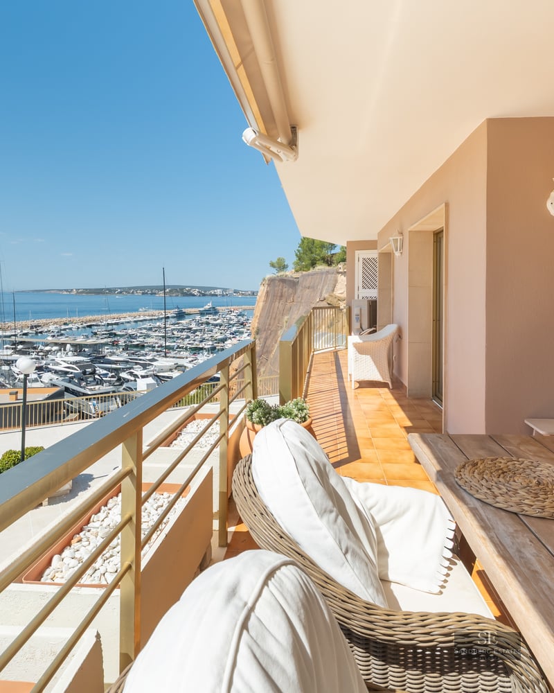 A sunlit balcony with a wooden dining table and wicker chairs overlooking a crowded marina and the blue ocean.
