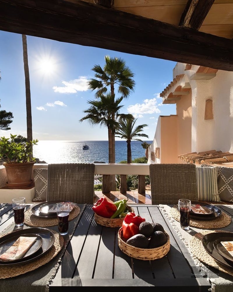 Outdoor dining table set on a sun-drenched terrace overlooking the Mediterranean sea and palm trees.