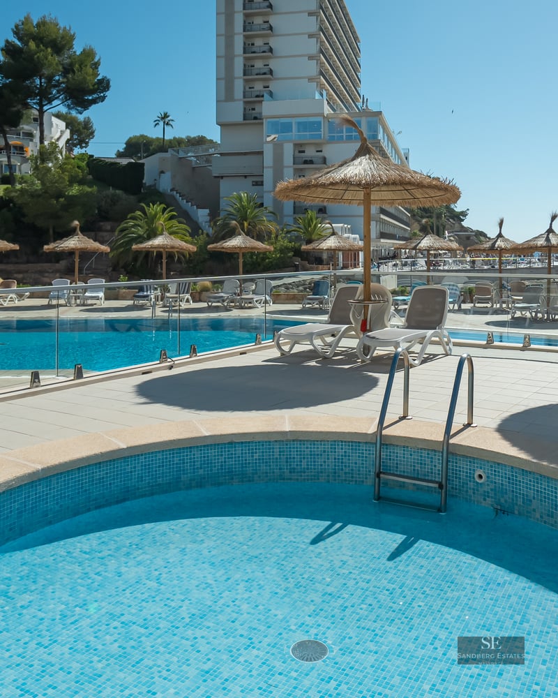 Bright outdoor pool area with straw umbrellas, white sun loungers, glass railings, and a view of the sea.