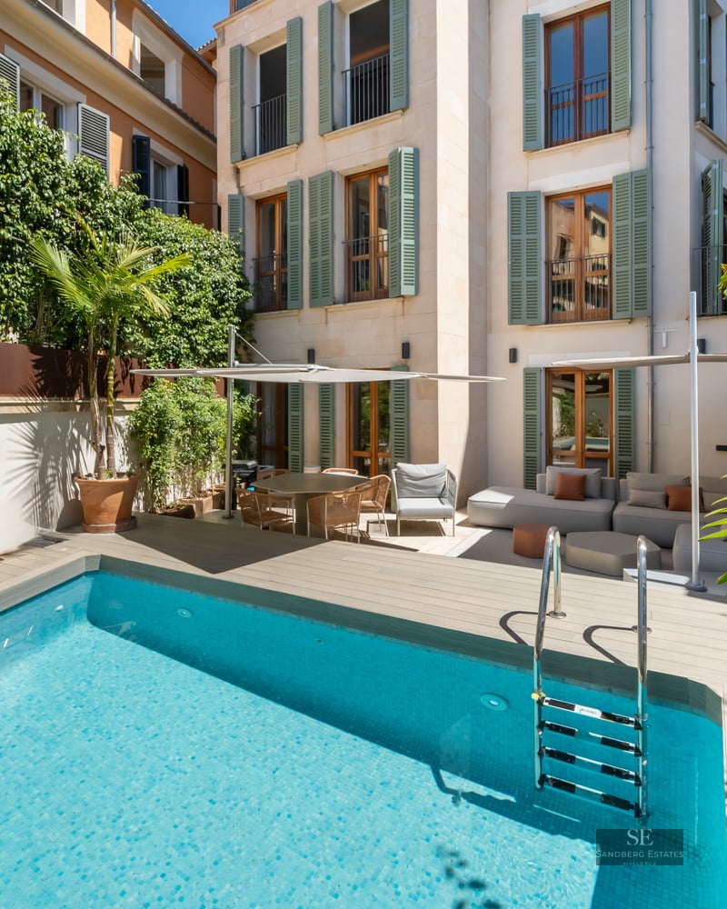 Light blue pool in a courtyard with wooden decking, outdoor sofas, and a building facade with green shutters.