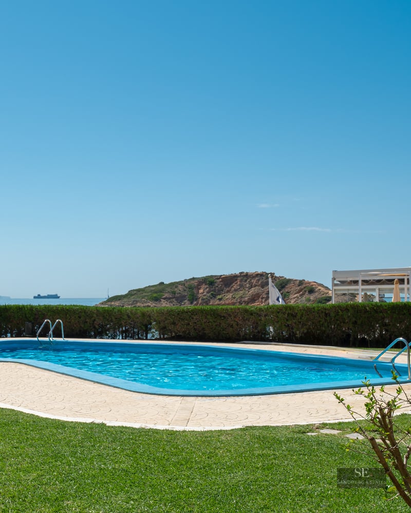 A blue curved swimming pool on a green lawn overlooking the Mediterranean sea under a clear sky.