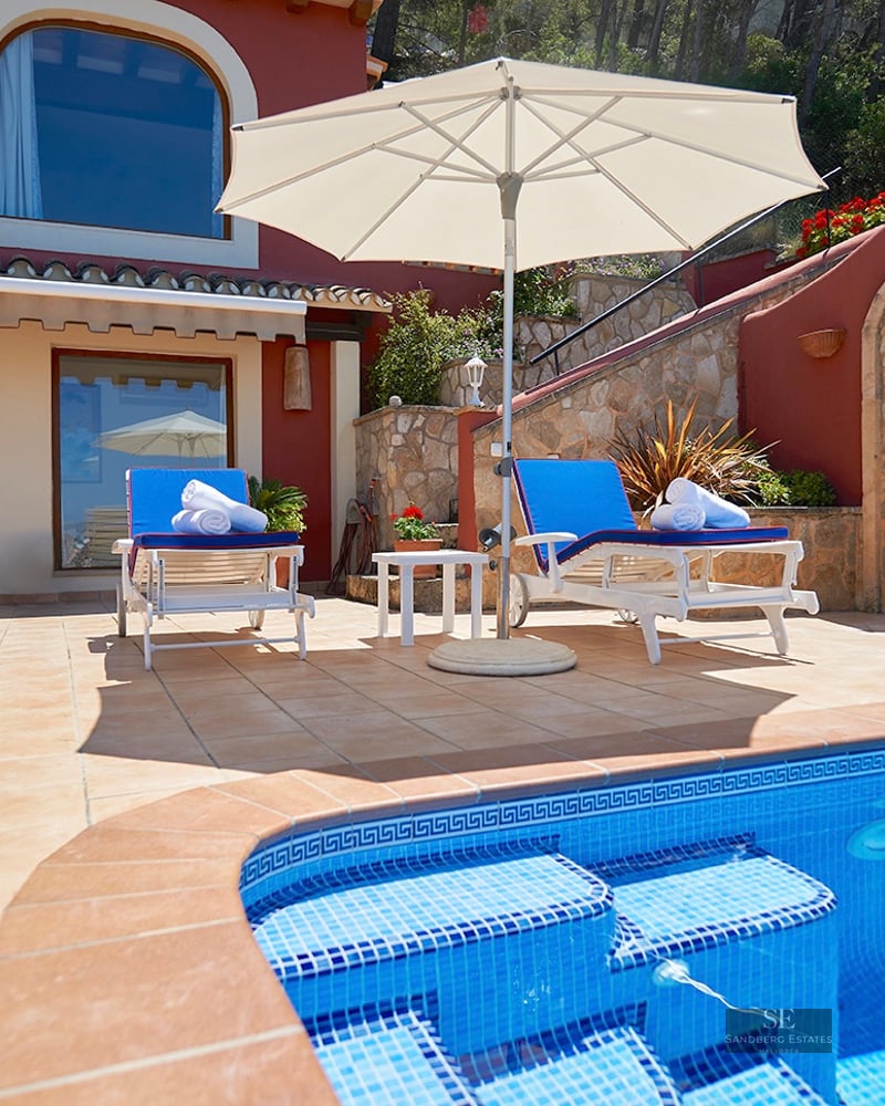 Blue tiled swimming pool with sun loungers and umbrella on a terracotta terrace next to a Mediterranean villa.