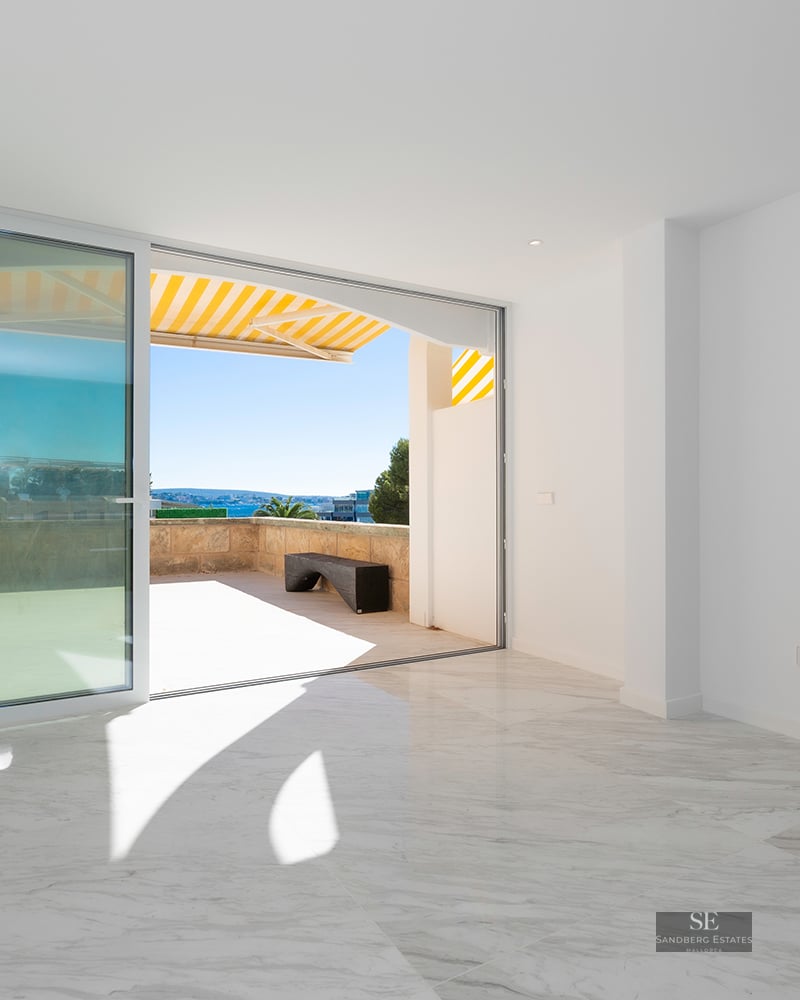 Bright empty room with white marble floors and a large sliding glass door opening to a sunny stone terrace.