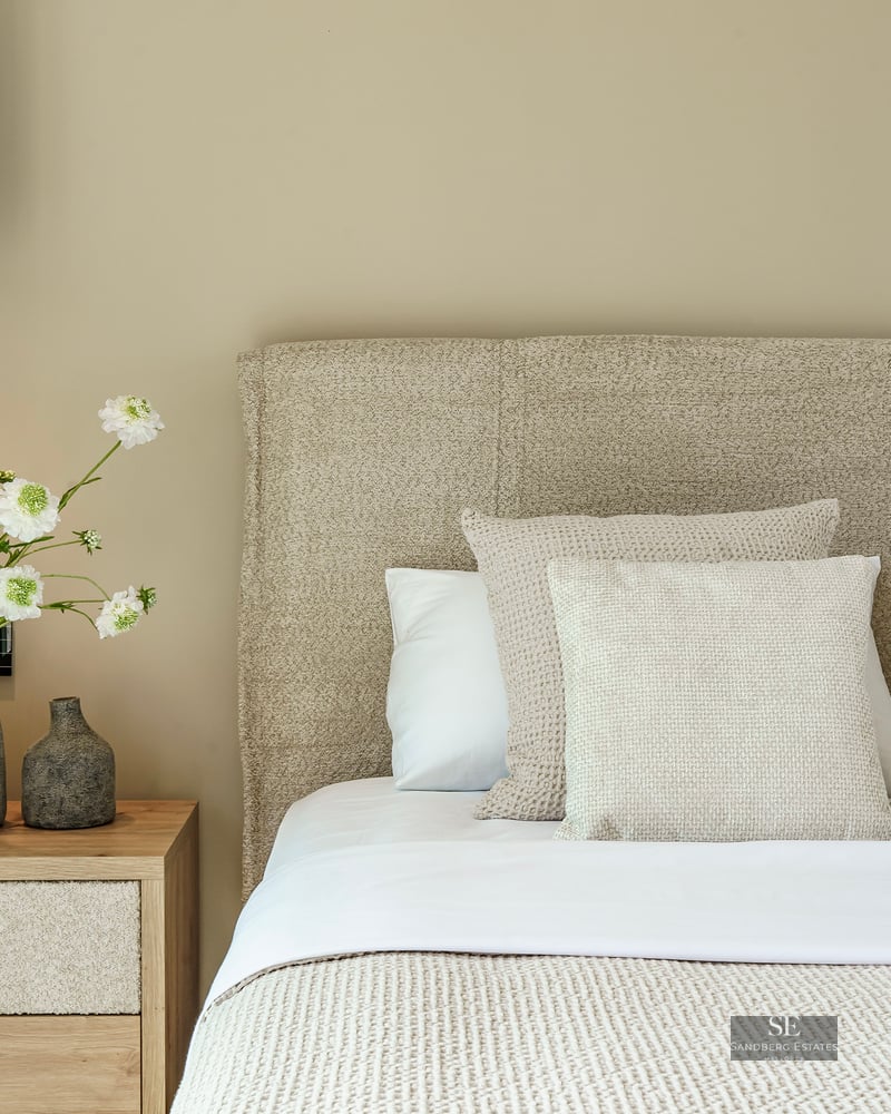 Close-up of a luxury bed with textured headboard, wooden nightstand, and decorative vases with white flowers.