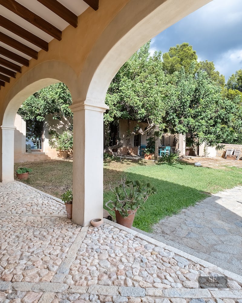 A traditional stone-arched porch with cobblestone flooring looking out onto a sunny garden with turquoise seating.