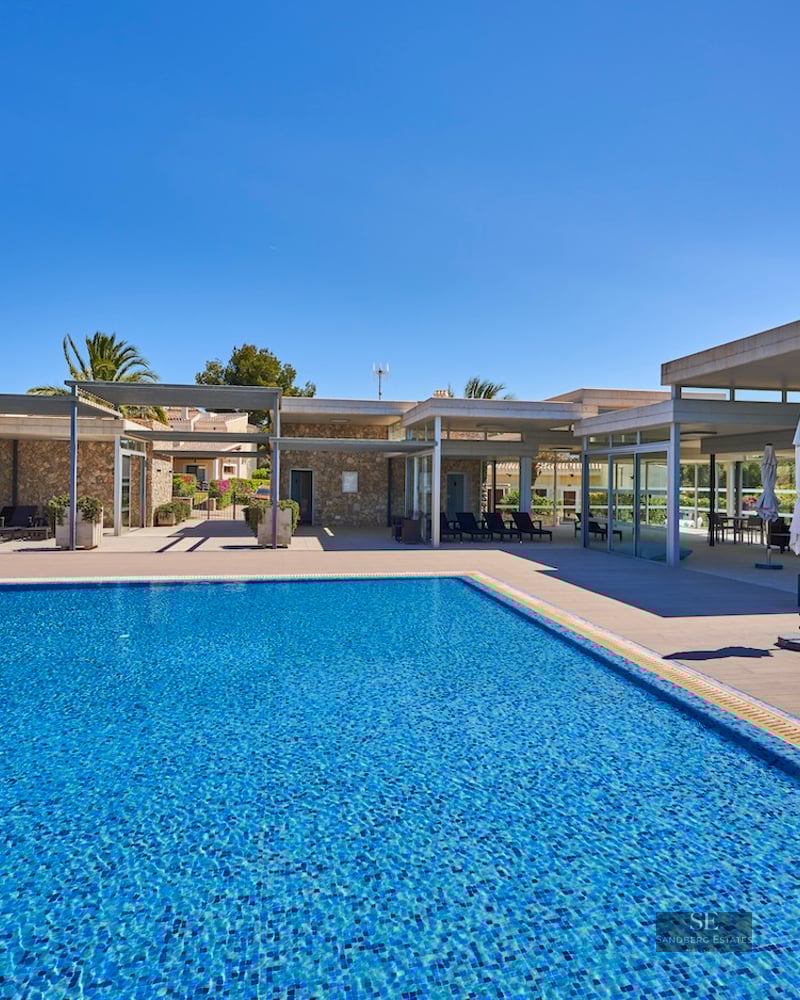 Large rectangular swimming pool with blue tiles next to a modern stone and glass villa under a clear blue sky.