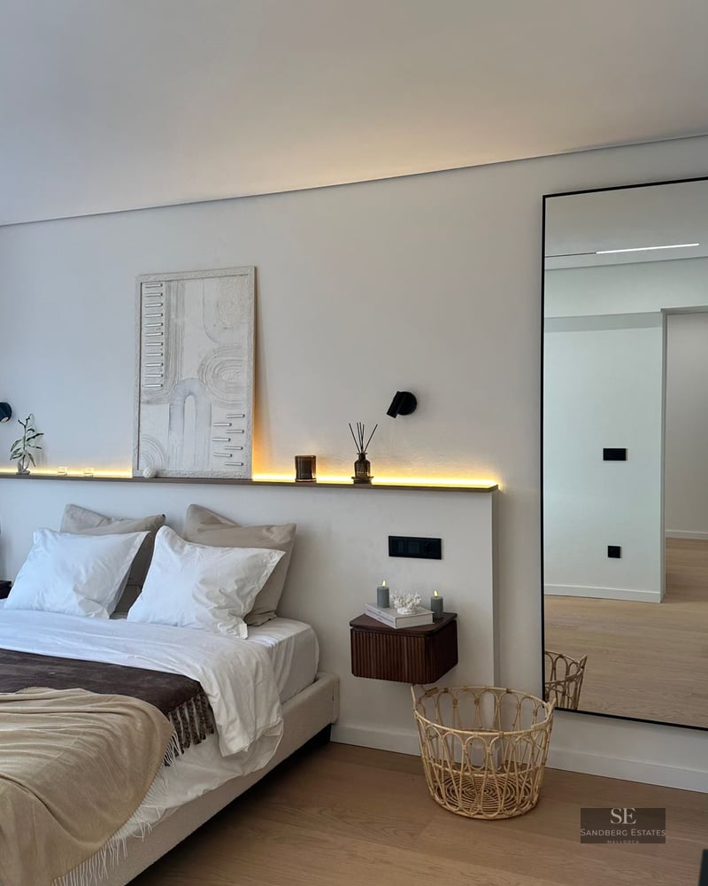 Modern bedroom featuring a white bed, LED backlighting, textured art, and a large mirror on light wood floors.