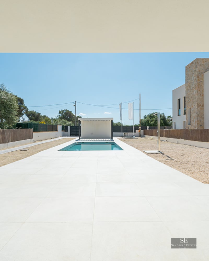 View from a covered porch toward a rectangular pool surrounded by white tiles and gravel in a modern villa setting.