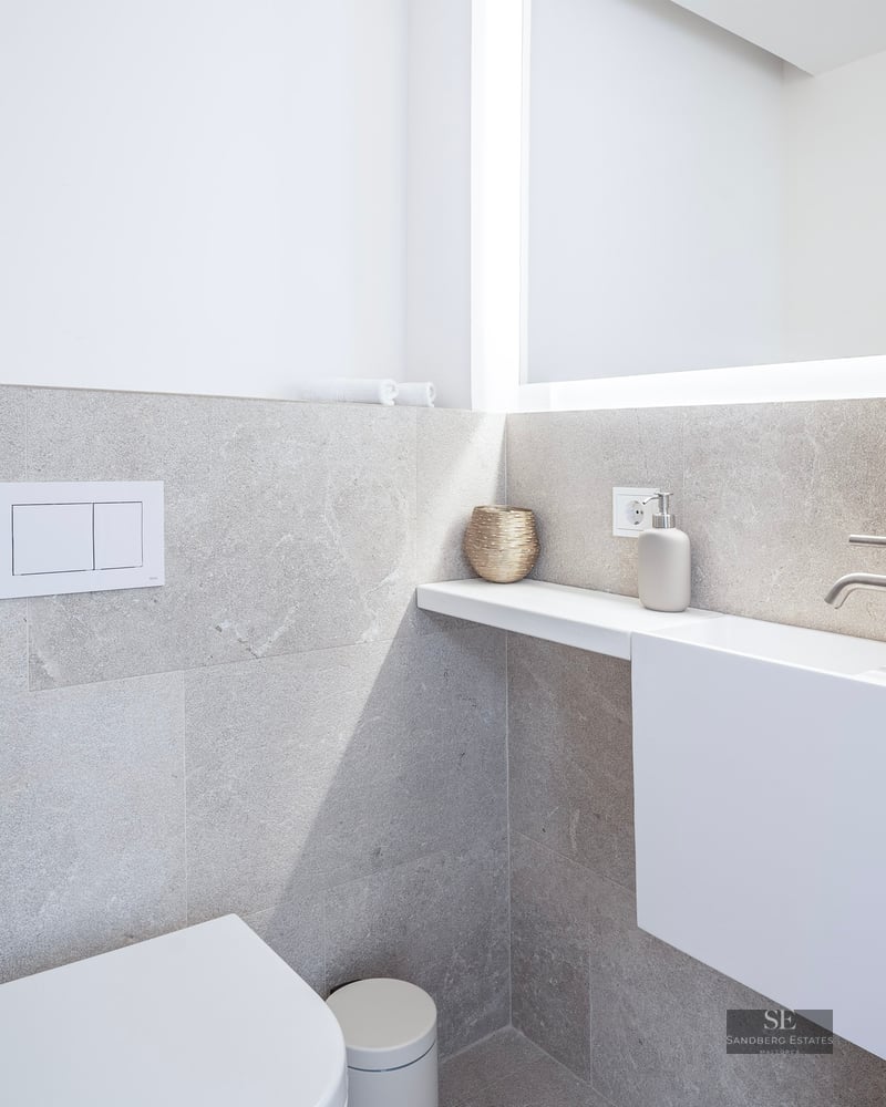 Salle de bain minimaliste avec WC blanc, petit lavabo suspendu et grand miroir rétroéclairé sur murs en pierre grise.