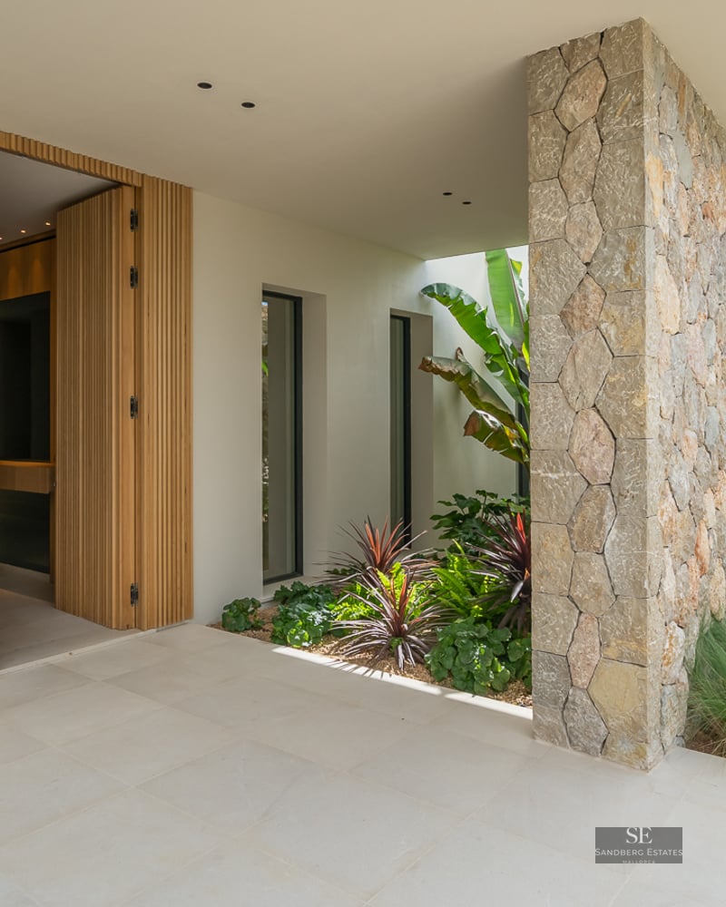 Modern home entrance featuring a slatted wood door, natural stone wall, and lush tropical landscaping.