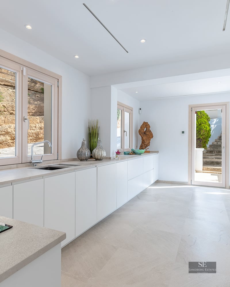 Bright modern white kitchen featuring stone countertops, large wood-framed windows, and integrated appliances.
