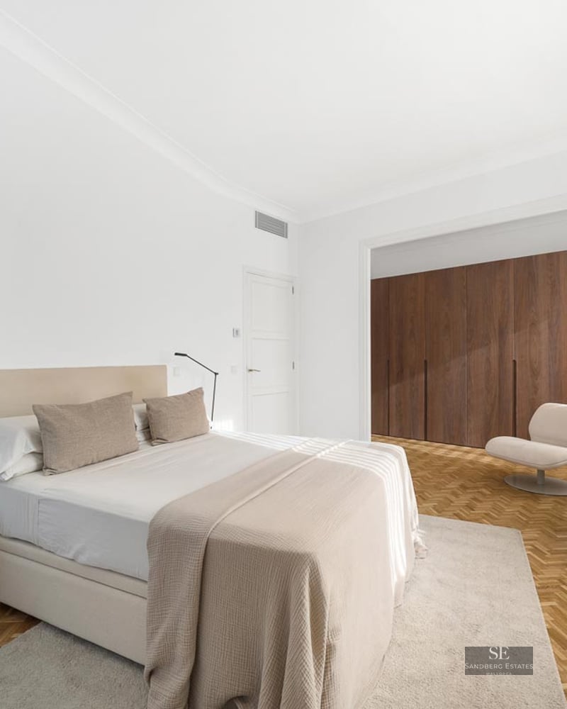Bright minimalist master bedroom featuring a large bed, herringbone wood floors, and floor-to-ceiling wooden wardrobes.