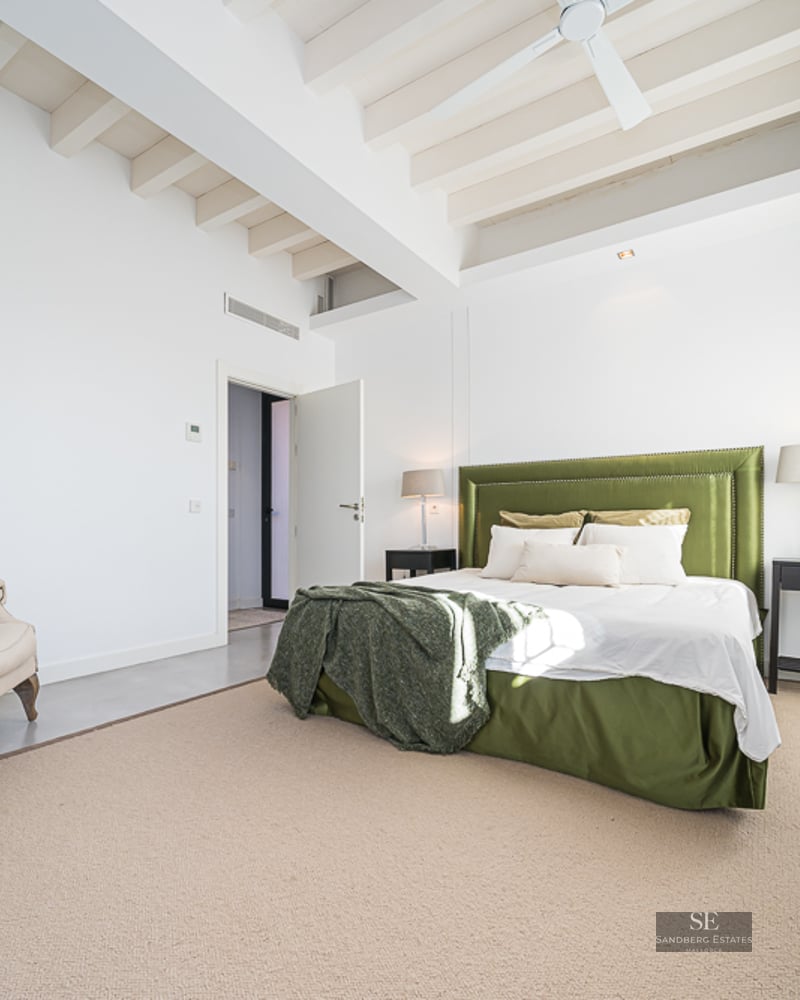 Bright bedroom with white exposed beams, olive green velvet bed, beige wingback chair, and large windows.