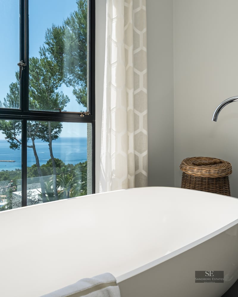 A white freestanding bathtub positioned in front of a large window overlooking the Mediterranean Sea and trees.