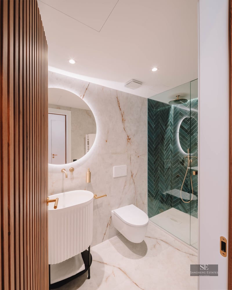 Modern bathroom featuring marble walls, a backlit circular mirror, gold fixtures, and green herringbone shower tiles.