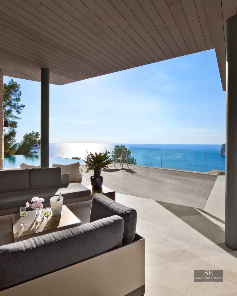 Outdoor lounge with grey sofas on a stone terrace overlooking an infinity pool and the Mediterranean Sea.