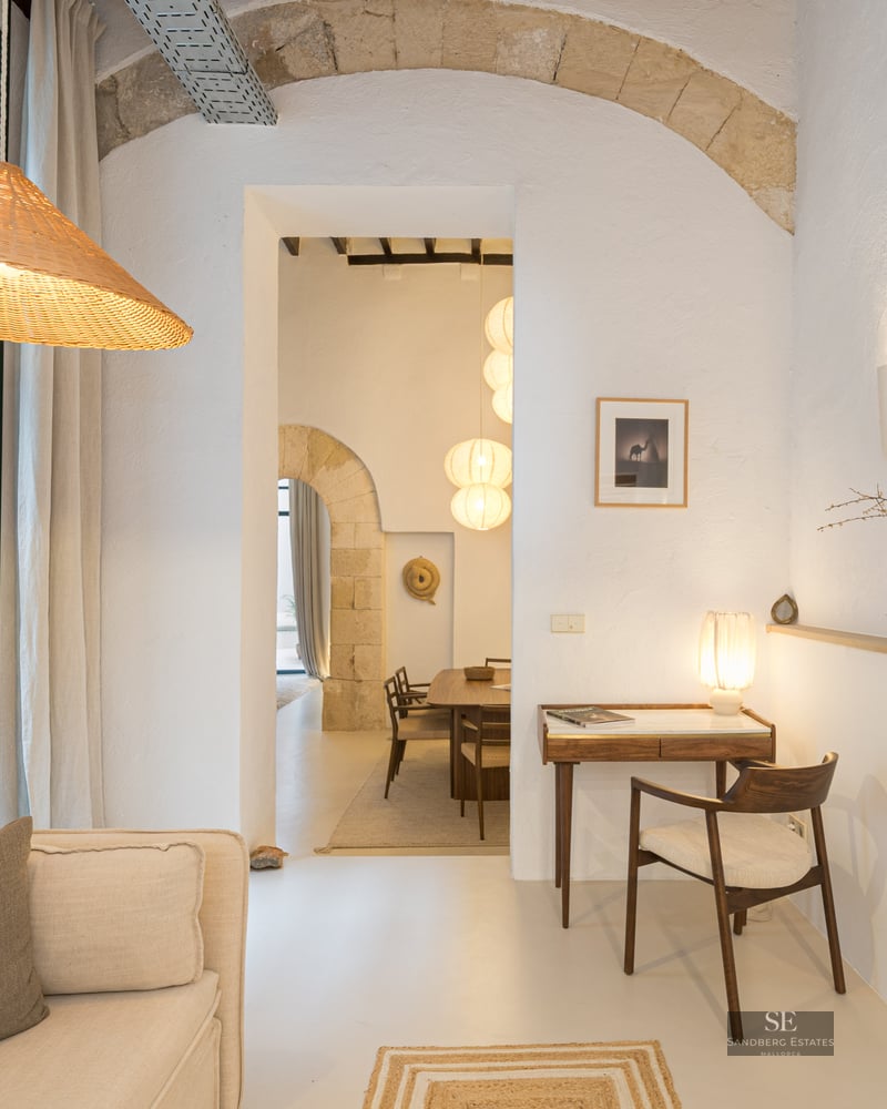 Salon blanc avec arches en pierre, suspension en osier, canapé et petit bureau en bois avec chaise.