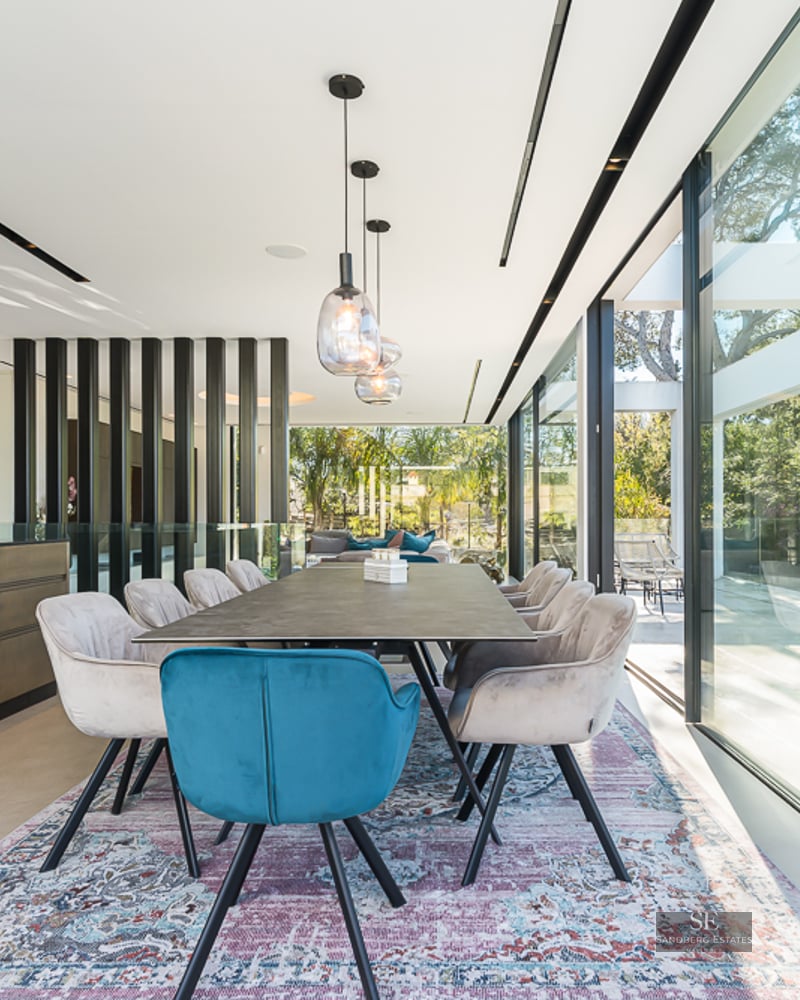 Bright dining room featuring a designer table, blue and beige velvet chairs, and floor-to-ceiling glass walls.