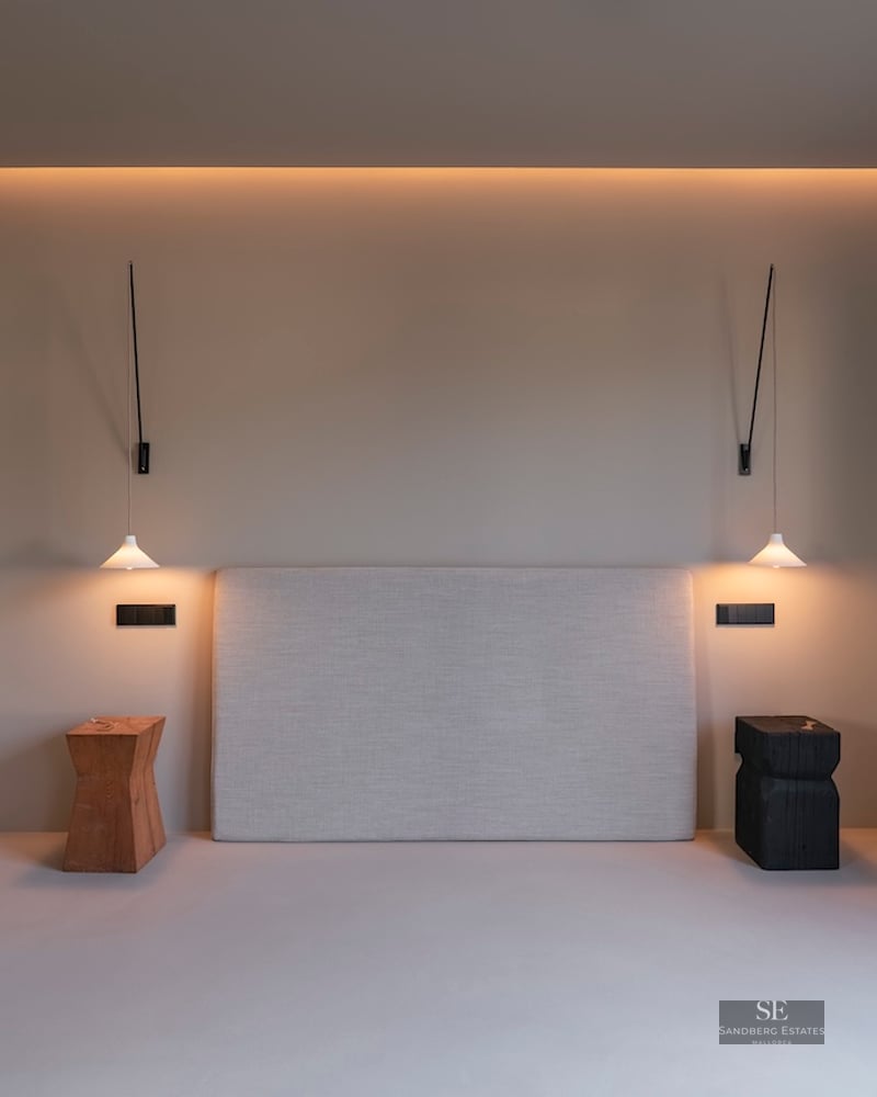 Tête de lit rembourrée entre des suspensions et des tabourets en bois sous un éclairage chaleureux et indirect.