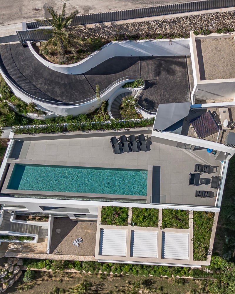Bird's eye drone view of a white modern villa with a rectangular pool, terrace furniture, and curved driveway.