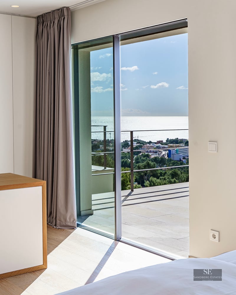 Bright modern bedroom with floor-to-ceiling sliding glass doors opening to a terrace overlooking the ocean.