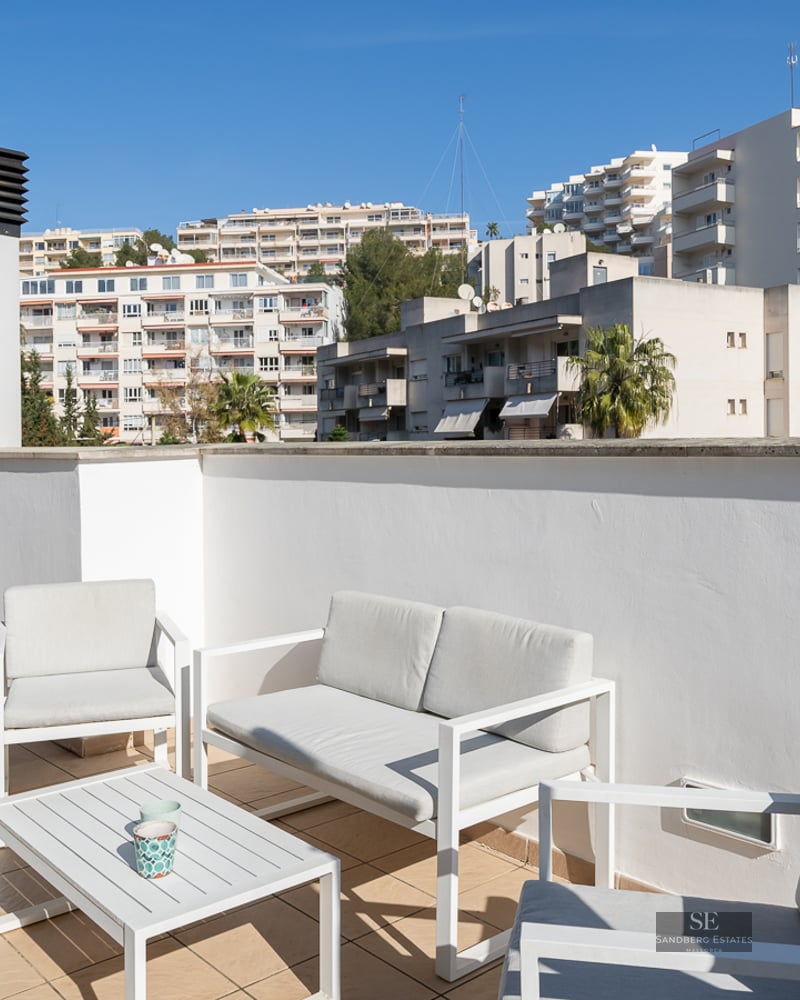Sunlit rooftop terrace with white modern furniture, tiled floor, and views of city buildings and mountains.