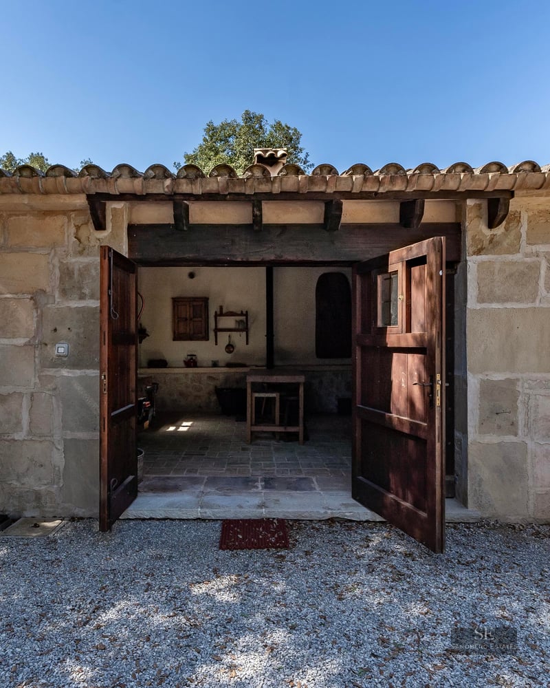 Small rustic stone building with open double wooden doors and a tiled roof on a sunny day.